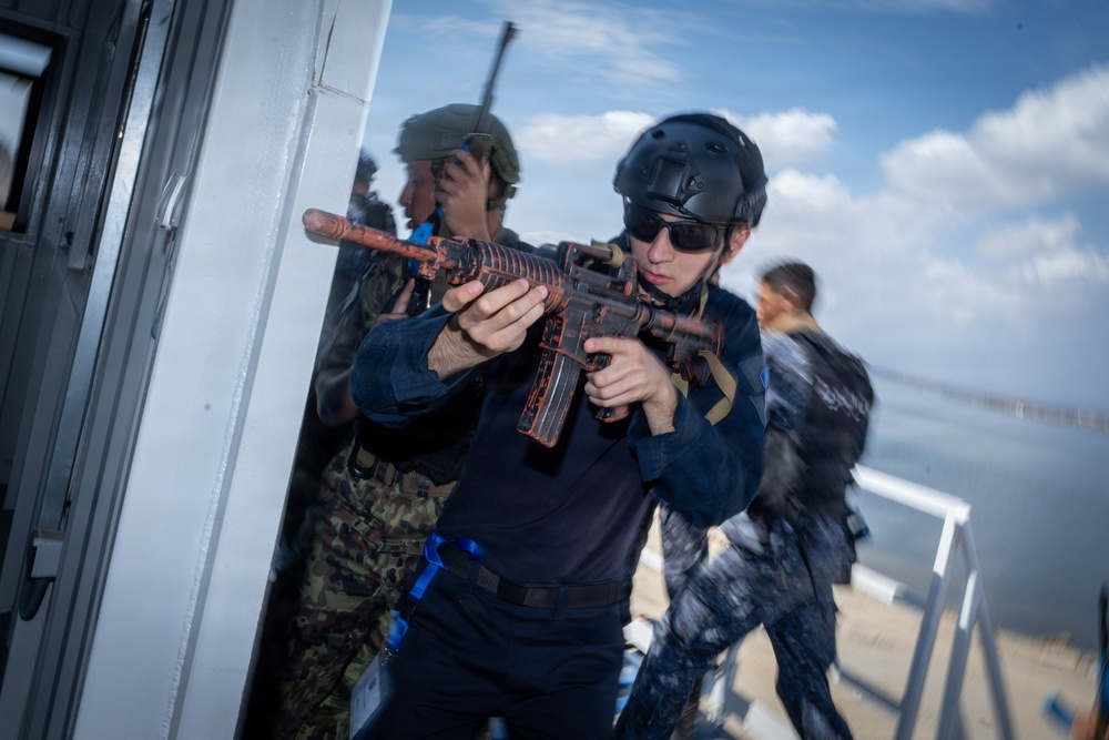 The image depicts what appears to be a military or law enforcement training exercise, with personnel in tactical gear conducting a building entry or clearing drill near a waterfront structure. The primary subject is wearing a black helmet, sunglasses, and dark uniform while aiming a rifle with a distinctive rust/orange-patterned finish. Additional personnel in mixed uniforms, including camouflage, are visible in the background.