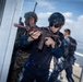 The image depicts what appears to be a military or law enforcement training exercise, with personnel in tactical gear conducting a building entry or clearing drill near a waterfront structure. The primary subject is wearing a black helmet, sunglasses, and dark uniform while aiming a rifle with a distinctive rust/orange-patterned finish. Additional personnel in mixed uniforms, including camouflage, are visible in the background.
