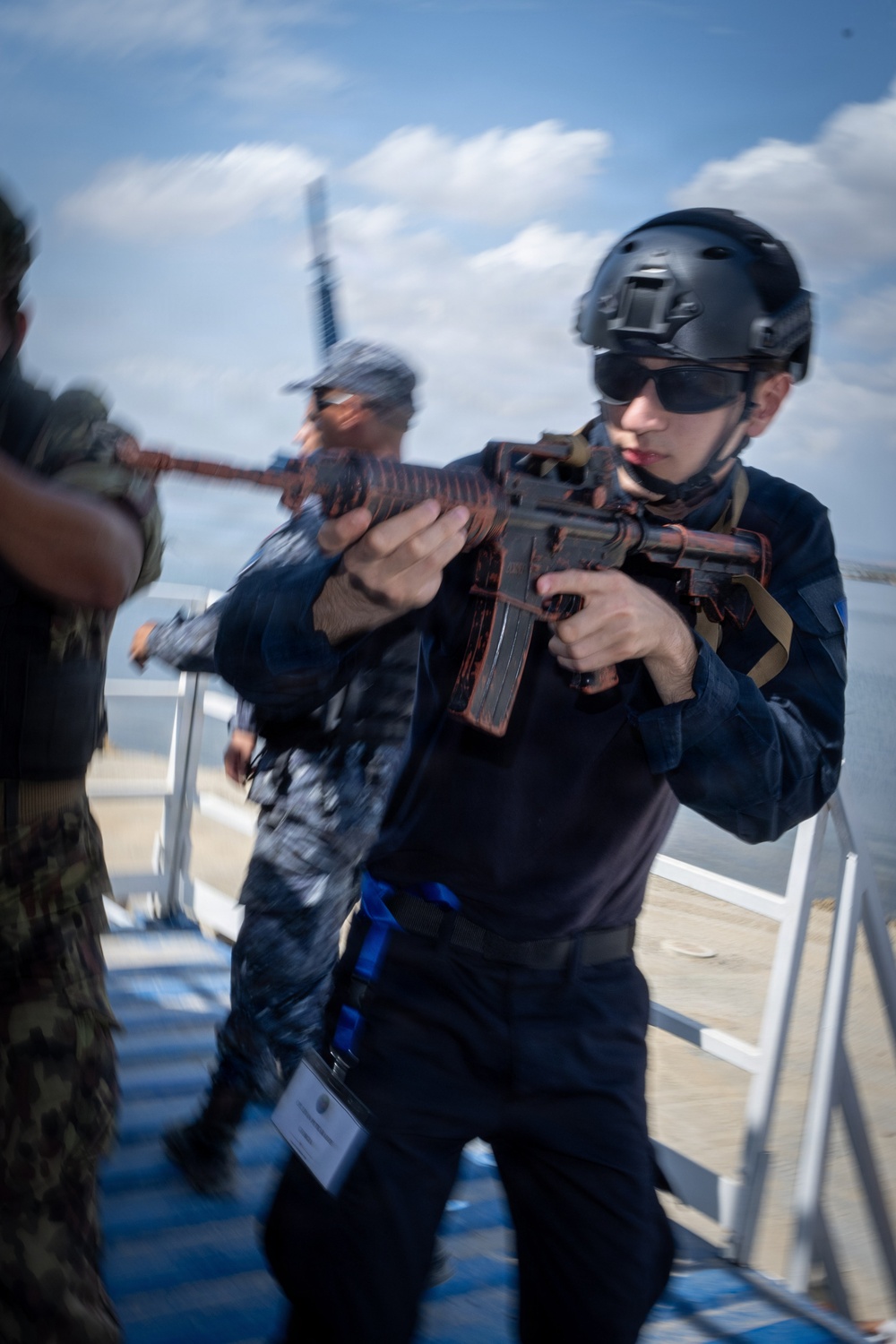 The image shows personnel conducting what appears to be a maritime security training exercise on the deck of a naval vessel. The primary subject is wearing all-black tactical gear including a helmet and sunglasses, aiming a weapon with orange/red training markings. Additional personnel in mixed military uniforms are visible in the background.