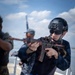The image shows personnel conducting what appears to be a maritime security training exercise on the deck of a naval vessel. The primary subject is wearing all-black tactical gear including a helmet and sunglasses, aiming a weapon with orange/red training markings. Additional personnel in mixed military uniforms are visible in the background.