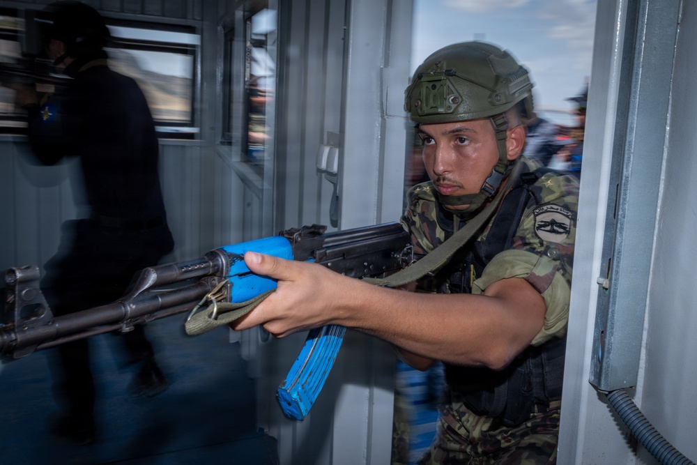 A soldier in camouflage uniform and tactical helmet is conducting what appears to be a military training exercise, aiming an AK-pattern rifle with blue training/MILES gear attachments through a doorway. The blue-colored magazine and handguard wrap indicate this is a training simulation rather than a live operation. A second figure in dark clothing is visible in the background, also appearing to participate in the exercise.