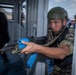 A soldier in camouflage uniform and tactical helmet is conducting what appears to be a military training exercise, aiming an AK-pattern rifle with blue training/MILES gear attachments through a doorway. The blue-colored magazine and handguard wrap indicate this is a training simulation rather than a live operation. A second figure in dark clothing is visible in the background, also appearing to participate in the exercise.