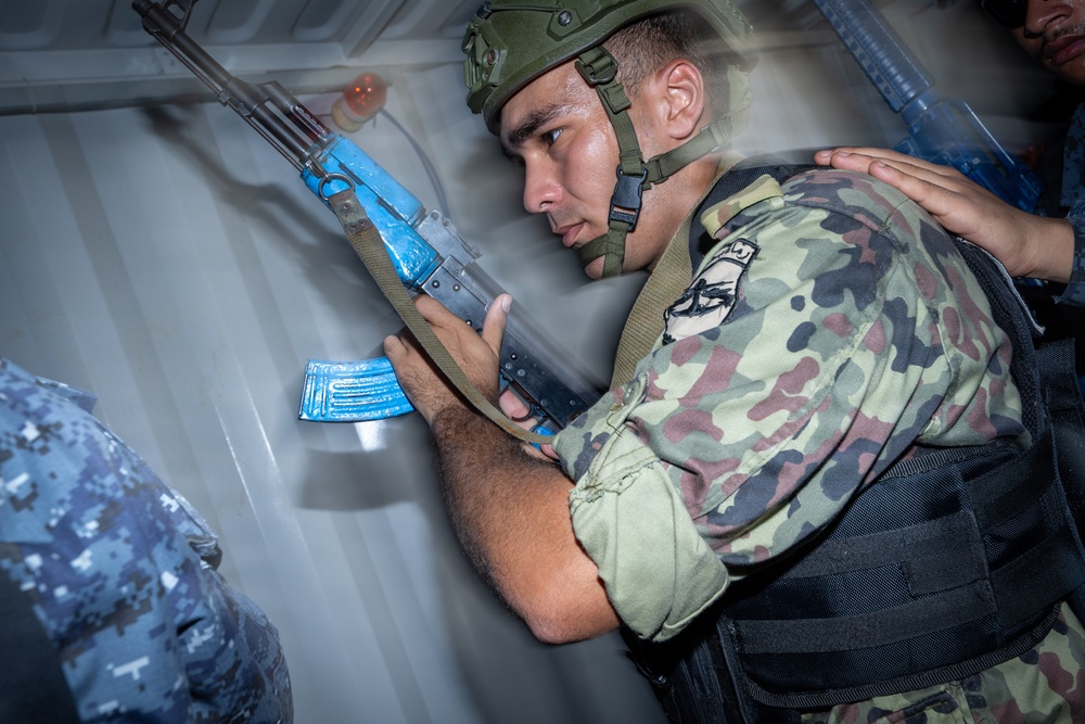 A soldier in camouflage uniform and helmet is conducting what appears to be a military training exercise, holding a rifle with blue-painted components indicating simunition or training ammunition. He is positioned against a metal wall in a tactical stance, with another person in blue digital camouflage visible nearby. The blue markings on the weapon and magazine are consistent with training/exercise equipment used to distinguish live weapons from training variants.