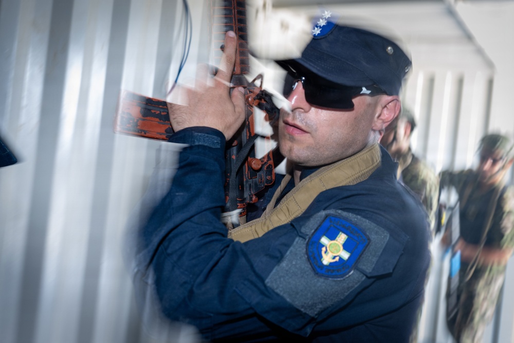 A uniformed security or law enforcement officer wearing a dark blue uniform with a shield-shaped patch is closely inspecting or breaching a metal container or door using a tool. The officer is wearing sunglasses and a dark cap with a star insignia. Additional personnel in camouflage uniforms are visible in the blurred background.