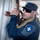 Allies, partners train together during Phoenix Express 2026 A uniformed security or law enforcement officer wearing a dark blue uniform with a shield-shaped patch is closely inspecting or breaching a metal container or door using a tool. The officer is wearing sunglasses and a dark cap with a star insignia. Additional personnel in camouflage uniforms are visible in the blurred background.