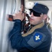 A uniformed security or law enforcement officer wearing a dark blue uniform with a shield-shaped patch is closely inspecting or breaching a metal container or door using a tool. The officer is wearing sunglasses and a dark cap with a star insignia. Additional personnel in camouflage uniforms are visible in the blurred background.