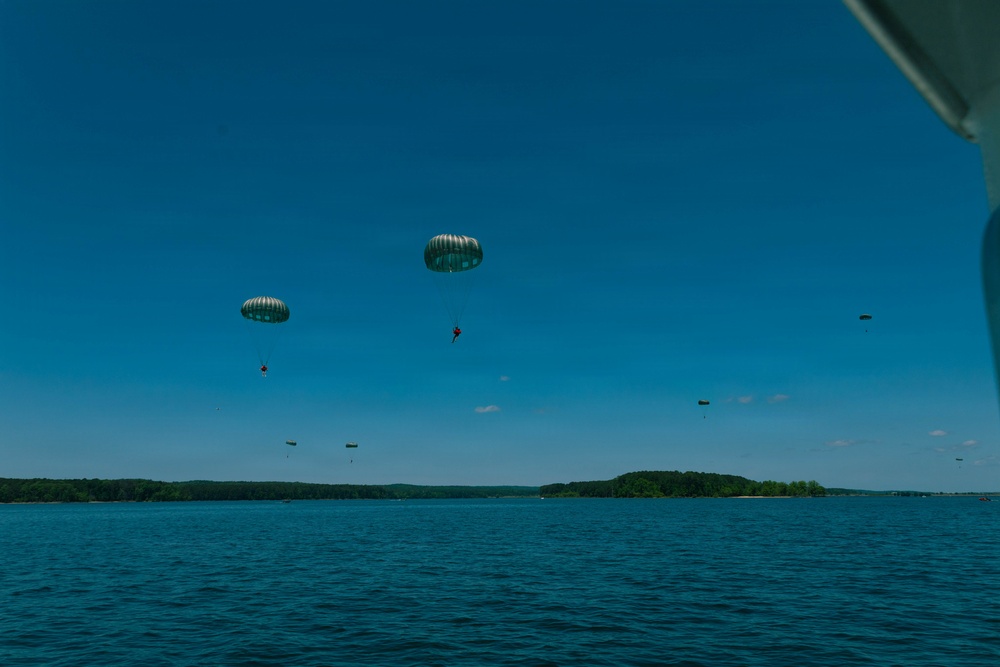 Multiple parachutists are descending over a large body of water under clear blue skies. At least two large, round military-style parachutes are clearly visible in the foreground, with several smaller parachutes visible in the background at greater distances. The scene appears to be a military or training water jump exercise, photographed from a boat or low aircraft.