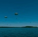 Multiple parachutists are descending over a large body of water under clear blue skies. At least two large, round military-style parachutes are clearly visible in the foreground, with several smaller parachutes visible in the background at greater distances. The scene appears to be a military or training water jump exercise, photographed from a boat or low aircraft.