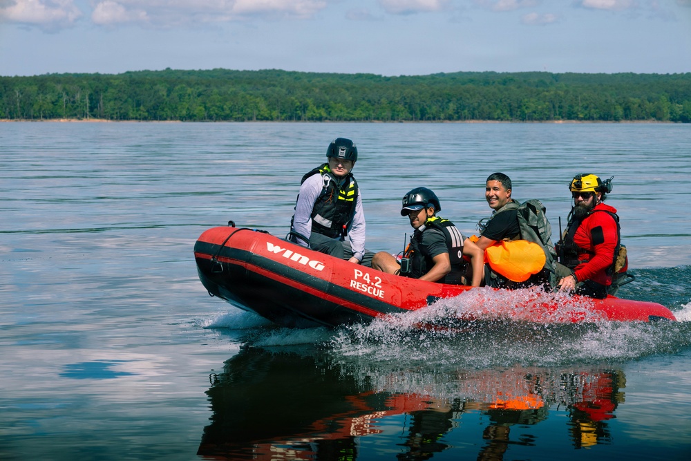 Four individuals are riding a red inflatable rescue boat labeled 'Wing P4.2 Rescue' across a calm body of water. The team appears to be conducting a water rescue operation or training exercise, with personnel wearing helmets, life vests, and tactical gear. A dense tree line is visible in the background under a partly cloudy sky.