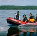 Four individuals are riding a red inflatable rescue boat labeled 'Wing P4.2 Rescue' across a calm body of water. The team appears to be conducting a water rescue operation or training exercise, with personnel wearing helmets, life vests, and tactical gear. A dense tree line is visible in the background under a partly cloudy sky.