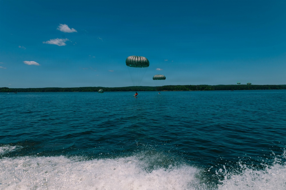 A scenic outdoor water scene showing parasailing activity over a large lake or bay on a clear sunny day. Two dome-shaped parachutes are visible in the air, with at least one person suspended beneath them above the water surface. The photo appears to be taken from a moving boat, as evidenced by the wake and foam visible in the foreground.