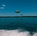 A scenic outdoor water scene showing parasailing activity over a large lake or bay on a clear sunny day. Two dome-shaped parachutes are visible in the air, with at least one person suspended beneath them above the water surface. The photo appears to be taken from a moving boat, as evidenced by the wake and foam visible in the foreground.