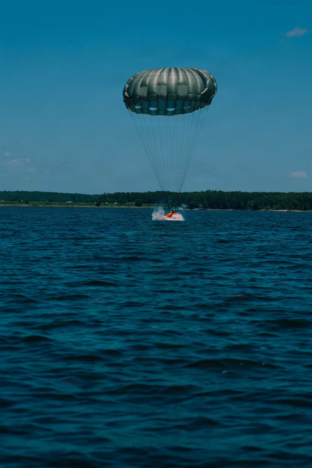 A parachute descent is captured at the moment of water landing on a large open body of water. A dark olive/gray round parachute canopy is fully deployed above a person or payload wearing or attached to orange equipment, creating a splash at the water's surface. A treeline and distant shoreline are visible on the horizon under a clear blue sky.