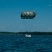 A parachute descent is captured at the moment of water landing on a large open body of water. A dark olive/gray round parachute canopy is fully deployed above a person or payload wearing or attached to orange equipment, creating a splash at the water's surface. A treeline and distant shoreline are visible on the horizon under a clear blue sky.