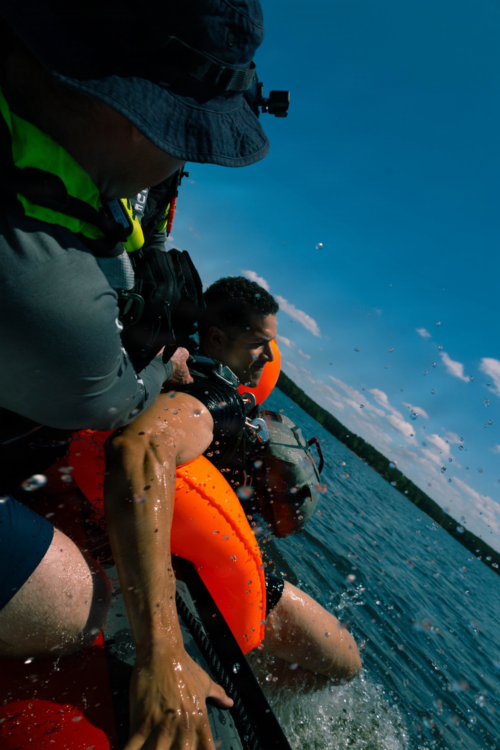 A water rescue or training exercise is taking place, with one person being pulled out of the water onto what appears to be a boat. The person being rescued is wearing an orange life vest and appears wet, while the rescuer wears a bucket hat with a mounted action camera and a bright green/yellow safety vest. Water droplets are visible mid-air, suggesting dynamic movement.