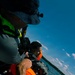 A water rescue or training exercise is taking place, with one person being pulled out of the water onto what appears to be a boat. The person being rescued is wearing an orange life vest and appears wet, while the rescuer wears a bucket hat with a mounted action camera and a bright green/yellow safety vest. Water droplets are visible mid-air, suggesting dynamic movement.