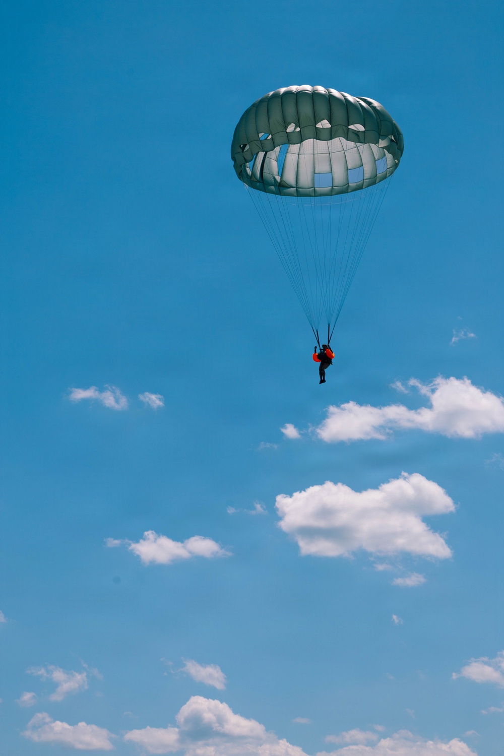 A single parachutist is descending through a clear blue sky with scattered white cumulus clouds. The parachute is a round, olive/grey-green military-style canopy with visible panels and suspension lines. The jumper appears to be wearing dark clothing with a distinctive red element on their torso.