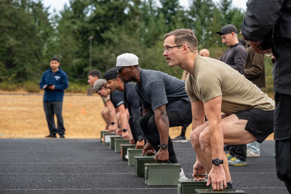 A group of individuals are positioned in a crouched starting stance on a paved track, each with their hands resting on olive-drab military ammunition cans. The scene appears to be a military or tactical fitness assessment, with observers standing nearby holding clipboards or notebooks. The setting is an outdoor track surrounded by coniferous trees under an overcast sky.
