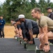 A group of individuals are positioned in a crouched starting stance on a paved track, each with their hands resting on olive-drab military ammunition cans. The scene appears to be a military or tactical fitness assessment, with observers standing nearby holding clipboards or notebooks. The setting is an outdoor track surrounded by coniferous trees under an overcast sky.