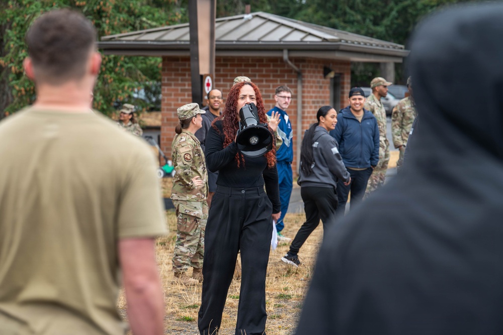 A woman with curly red hair dressed in all black is speaking into a megaphone/bullhorn while addressing a group of people outdoors. The crowd includes individuals in military camouflage uniforms as well as civilians in casual athletic wear. The setting appears to be a military installation or base, indicated by the brick building with a red cross symbol in the background.