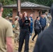 A woman with curly red hair dressed in all black is speaking into a megaphone/bullhorn while addressing a group of people outdoors. The crowd includes individuals in military camouflage uniforms as well as civilians in casual athletic wear. The setting appears to be a military installation or base, indicated by the brick building with a red cross symbol in the background.