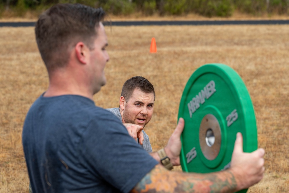 Two men are engaged in an outdoor fitness activity on a dry grass field. One man in the foreground holds a green 25-pound bumper plate weight, while a second man in the background appears to be in a low position, possibly resting or preparing for an exercise. An orange traffic cone is visible in the background, suggesting a marked course or training area.