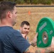Two men are engaged in an outdoor fitness activity on a dry grass field. One man in the foreground holds a green 25-pound bumper plate weight, while a second man in the background appears to be in a low position, possibly resting or preparing for an exercise. An orange traffic cone is visible in the background, suggesting a marked course or training area.