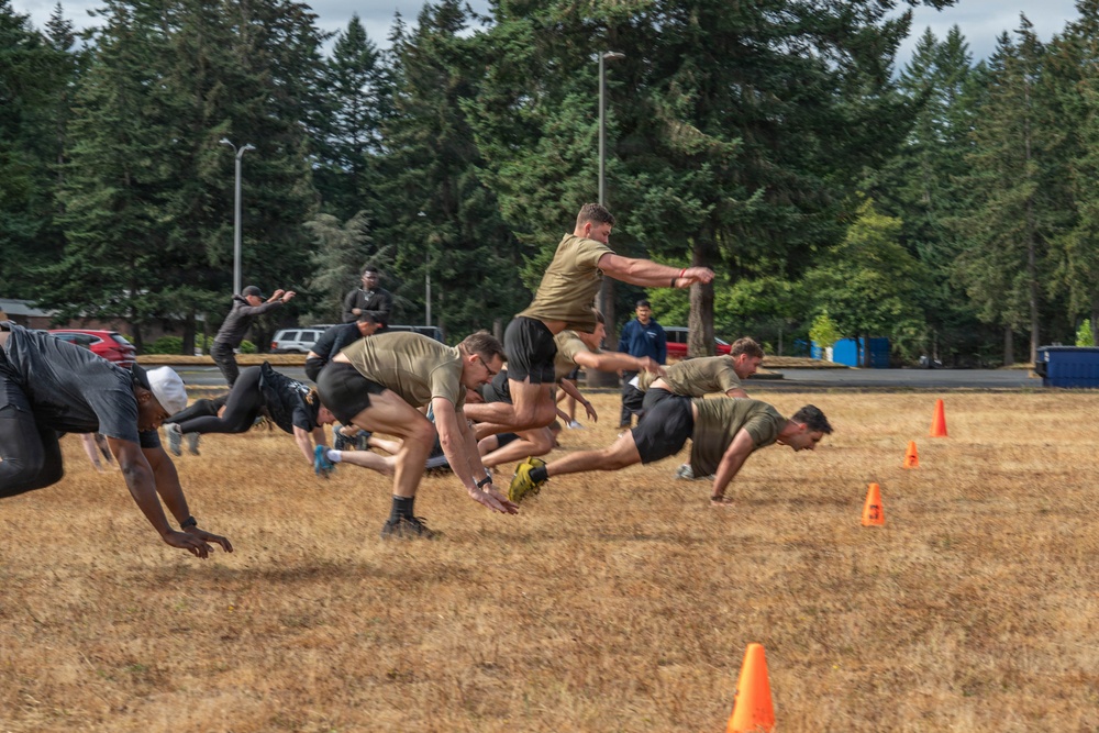 A group of individuals in military-style athletic wear are performing a burpee or dive-bomber exercise drill on a dry grass field. The participants appear to be in mid-motion, lunging or diving forward in a synchronized manner, suggesting a structured physical training exercise. Orange traffic cones mark the field boundaries, and the setting appears to be a military base or institutional grounds.