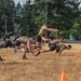 A group of individuals in military-style athletic wear are performing a burpee or dive-bomber exercise drill on a dry grass field. The participants appear to be in mid-motion, lunging or diving forward in a synchronized manner, suggesting a structured physical training exercise. Orange traffic cones mark the field boundaries, and the setting appears to be a military base or institutional grounds.