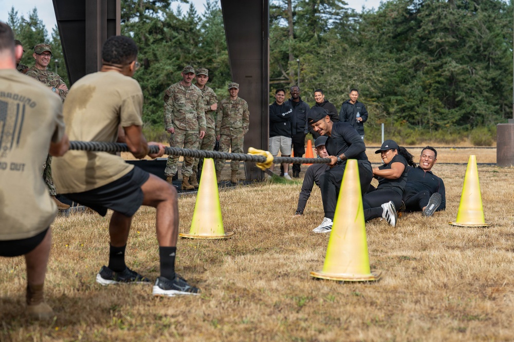 A tug-of-war competition is taking place on a dry grass field, with participants divided into two teams pulling a thick rope. Yellow traffic cones mark the boundary line between the competing teams. Military personnel in camouflage uniforms and civilians in black athletic wear observe and participate in the event.