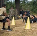 A tug-of-war competition is taking place on a dry grass field, with participants divided into two teams pulling a thick rope. Yellow traffic cones mark the boundary line between the competing teams. Military personnel in camouflage uniforms and civilians in black athletic wear observe and participate in the event.