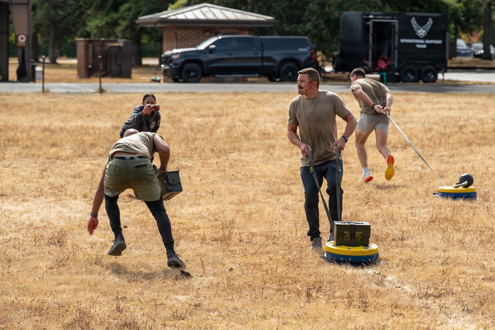 A group of individuals are participating in an outdoor fitness or military-style training exercise on a dry, grassy field. Participants are performing various physical tasks including carrying ammunition cans and dragging weighted sleds. A black trailer with a US Air Force insignia is visible in the background, suggesting a military or Air Force-affiliated event.