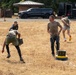 A group of individuals are participating in an outdoor fitness or military-style training exercise on a dry, grassy field. Participants are performing various physical tasks including carrying ammunition cans and dragging weighted sleds. A black trailer with a US Air Force insignia is visible in the background, suggesting a military or Air Force-affiliated event.