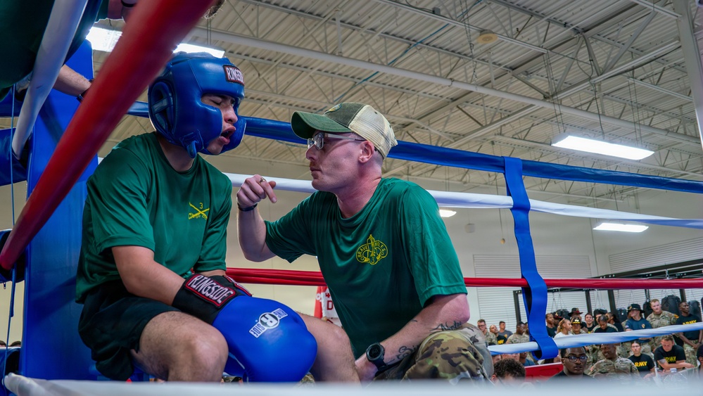 A boxing coach or cornerman is giving instructions to a boxer during a break between rounds in a military boxing event. Both individuals are wearing matching green t-shirts with what appears to be a military unit insignia, suggesting this is an Army-sponsored boxing competition. A large audience of military personnel in uniform can be seen in the background.