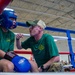 A boxing coach or cornerman is giving instructions to a boxer during a break between rounds in a military boxing event. Both individuals are wearing matching green t-shirts with what appears to be a military unit insignia, suggesting this is an Army-sponsored boxing competition. A large audience of military personnel in uniform can be seen in the background.