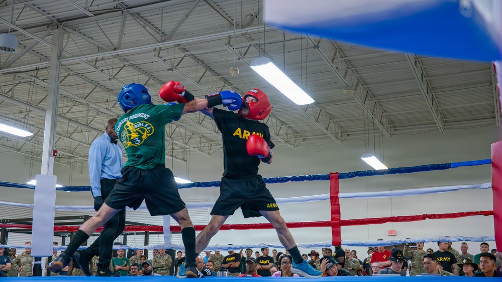 Two U.S. Army soldiers are competing in a boxing match inside an indoor facility, wearing protective headgear and gloves. One boxer wears a green 'Cavalry Regiment' shirt with a blue helmet, while the other wears a black 'ARMY' shirt with a red helmet. A large crowd of military personnel in camouflage uniforms watches from ringside, with a referee visible in the background.