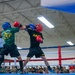 Two U.S. Army soldiers are competing in a boxing match inside an indoor facility, wearing protective headgear and gloves. One boxer wears a green 'Cavalry Regiment' shirt with a blue helmet, while the other wears a black 'ARMY' shirt with a red helmet. A large crowd of military personnel in camouflage uniforms watches from ringside, with a referee visible in the background.