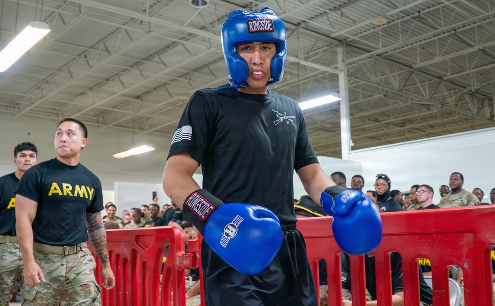 A U.S. Army soldier wearing blue Ringside boxing headgear and gloves stands in a combat sports area surrounded by red barriers, with a crowd of fellow soldiers watching in the background. The setting appears to be an indoor military gymnasium or training facility hosting a boxing or combatives event. The soldier is in a focused, ready stance suggesting active participation in a sparring or competition event.
