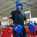 A U.S. Army soldier wearing blue Ringside boxing headgear and gloves stands in a combat sports area surrounded by red barriers, with a crowd of fellow soldiers watching in the background. The setting appears to be an indoor military gymnasium or training facility hosting a boxing or combatives event. The soldier is in a focused, ready stance suggesting active participation in a sparring or competition event.