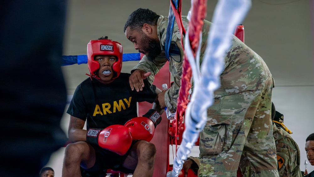 A military boxing event showing a boxer in a corner between rounds. The seated boxer wears an 'ARMY' t-shirt, red Ringside headgear, red Ringside boxing gloves, and a mouthguard, while a cornerman in U.S. Army OCP (camouflage) uniform attends to them. The setting appears to be an indoor military boxing competition with ring ropes visible in the foreground.