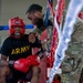 A military boxing event showing a boxer in a corner between rounds. The seated boxer wears an 'ARMY' t-shirt, red Ringside headgear, red Ringside boxing gloves, and a mouthguard, while a cornerman in U.S. Army OCP (camouflage) uniform attends to them. The setting appears to be an indoor military boxing competition with ring ropes visible in the foreground.