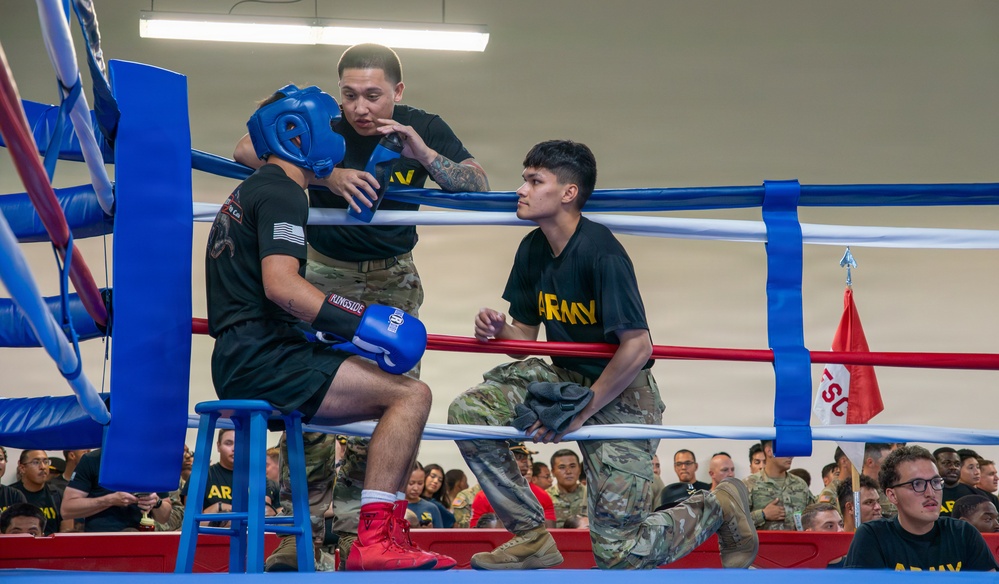 A military boxing event is taking place in a ring with blue and red ropes. A boxer wearing a blue headguard, blue Ringside gloves, and red boxing shoes is seated on a stool in the corner, receiving coaching from two individuals wearing U.S. Army t-shirts and camouflage pants during a between-rounds break. A large crowd of uniformed military personnel watches from the background.