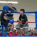 A military boxing event is taking place in a ring with blue and red ropes. A boxer wearing a blue headguard, blue Ringside gloves, and red boxing shoes is seated on a stool in the corner, receiving coaching from two individuals wearing U.S. Army t-shirts and camouflage pants during a between-rounds break. A large crowd of uniformed military personnel watches from the background.