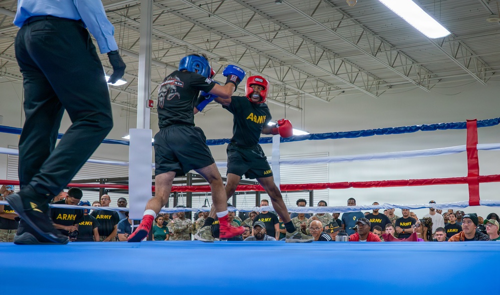 Two U.S. Army soldiers are competing in a boxing match inside an indoor facility, both wearing protective headgear and gloves. A referee in a light blue shirt and black pants oversees the bout from the left side of the ring. A large crowd of military personnel and civilians watches from ringside.