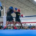 Two U.S. Army soldiers are competing in a boxing match inside an indoor facility, both wearing protective headgear and gloves. A referee in a light blue shirt and black pants oversees the bout from the left side of the ring. A large crowd of military personnel and civilians watches from ringside.