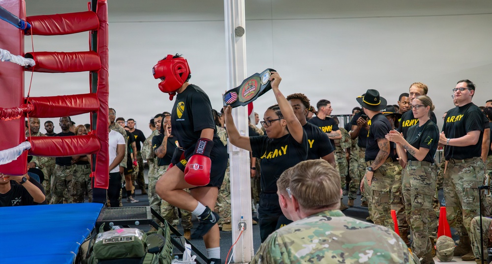 A military boxing event is taking place in an indoor facility, where a boxer wearing red headgear and red gloves is being presented with a championship belt by a person in an Army black t-shirt. The event is attended by a large crowd of U.S. Army personnel in both camouflage uniforms and black Army PT shirts. The atmosphere appears celebratory, suggesting this is a post-match award ceremony.