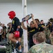 A military boxing event is taking place in an indoor facility, where a boxer wearing red headgear and red gloves is being presented with a championship belt by a person in an Army black t-shirt. The event is attended by a large crowd of U.S. Army personnel in both camouflage uniforms and black Army PT shirts. The atmosphere appears celebratory, suggesting this is a post-match award ceremony.
