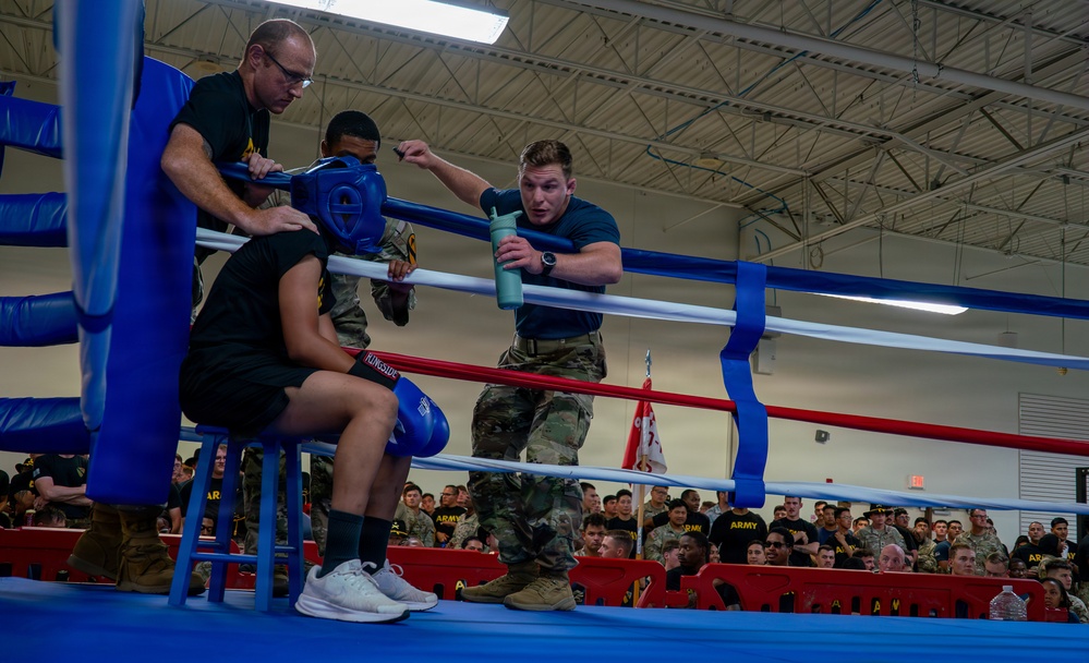 A military boxing event is taking place inside an indoor gymnasium facility. A boxer is seated in the corner of the ring between rounds while cornermen attend to him, adjusting his headgear and providing water. A large audience of military personnel in Army uniforms watches from the stands.