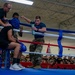 A military boxing event is taking place inside an indoor gymnasium facility. A boxer is seated in the corner of the ring between rounds while cornermen attend to him, adjusting his headgear and providing water. A large audience of military personnel in Army uniforms watches from the stands.