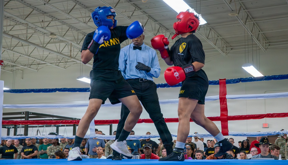 Two U.S. Army soldiers are competing in a boxing match inside what appears to be a military facility or gymnasium. A referee dressed in a light blue shirt and bow tie oversees the bout as one boxer in blue gear throws a punch at the opponent in red gear. A large crowd of military personnel and spectators watches from the background.